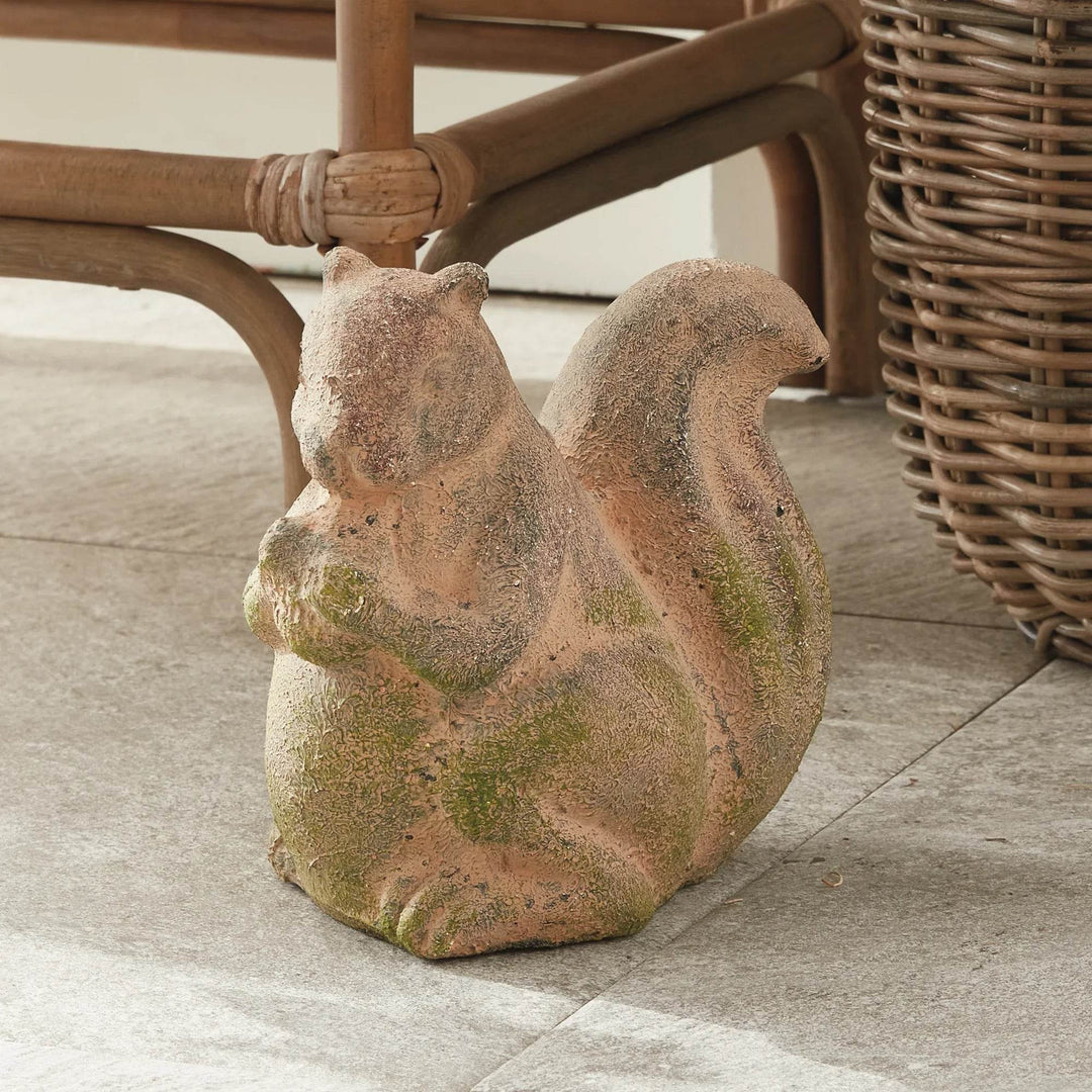Weathered terracotta squirrel sculpture with mossy finish, placed outdoors next to wicker furniture and natural stone flooring.