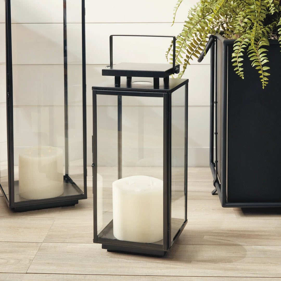 Small black outdoor lantern with glass panels and pillar candle, styled on porch near greenery and wicker chair – Transitional