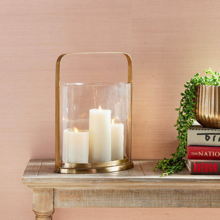 Brass and glass hurricane candle holder with pillar candles displayed on wooden console beside books and planter