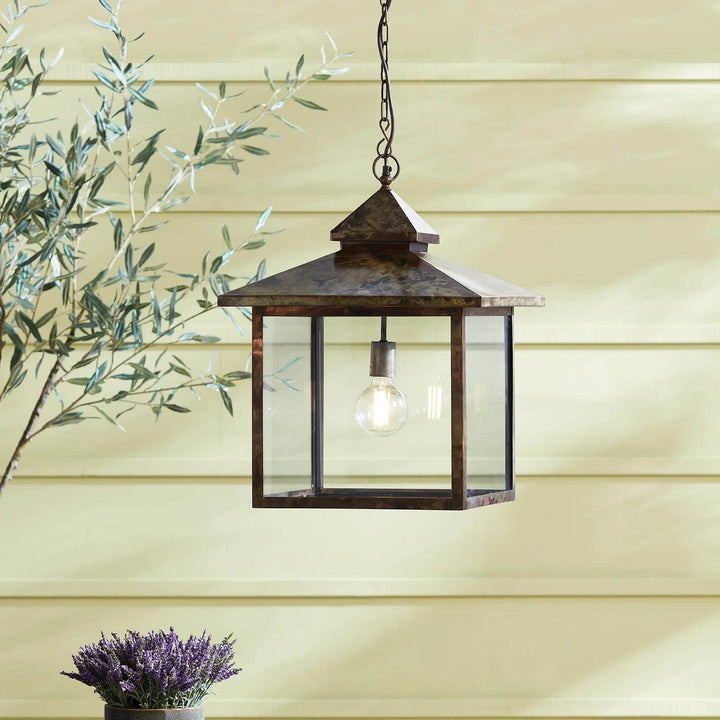 Heritage Rustic hanging lantern displayed outdoors with greenery and warm light against soft yellow siding.