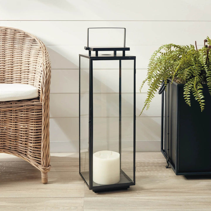 Large rectangular black outdoor lantern with glass sides, shown near wicker chair and greenery for porch or entryway – Transitional