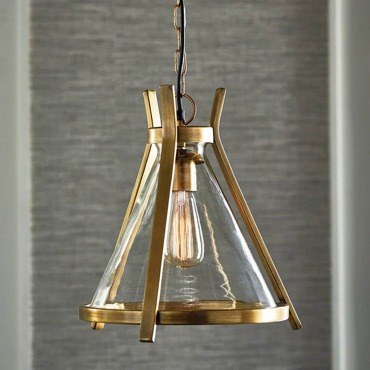 Modern industrial pendant light with antique brass frame and clear glass shade, shown hanging against a textured gray backdrop