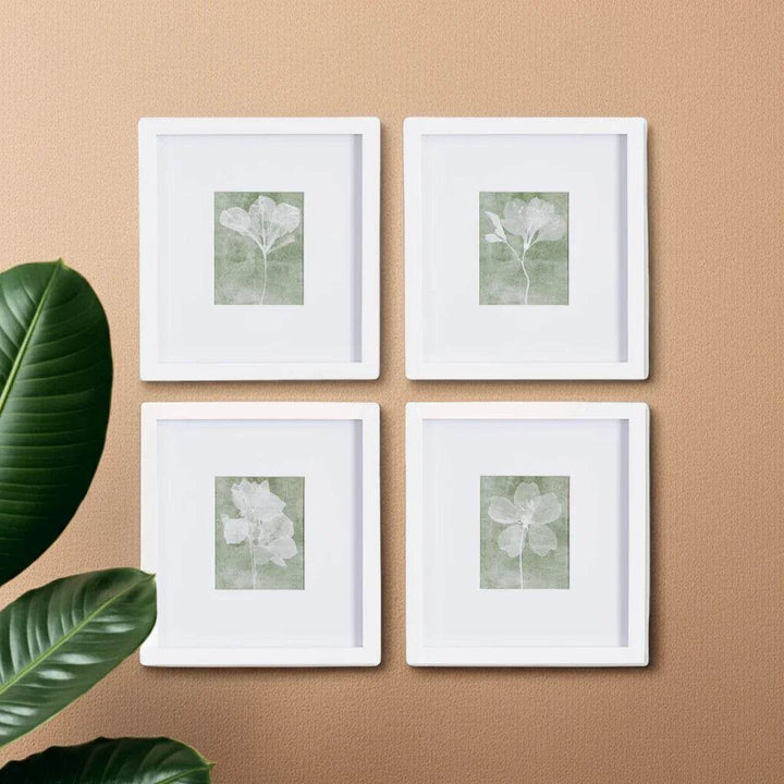 Soft green flower prints in white frames on a peach-toned wall with leafy accent plant