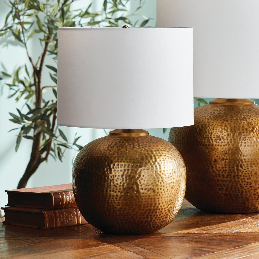 Enhance any room with the elegant style of Tara Gold &amp; White Table Lamp Small. The warm brass round base exudes a luxurious feel, while the crisp white linen drum shade offers a gentle radiance. Crafted from hammered iron and featuring unique handmade elements, this lamp is the perfect addition to any modern space.