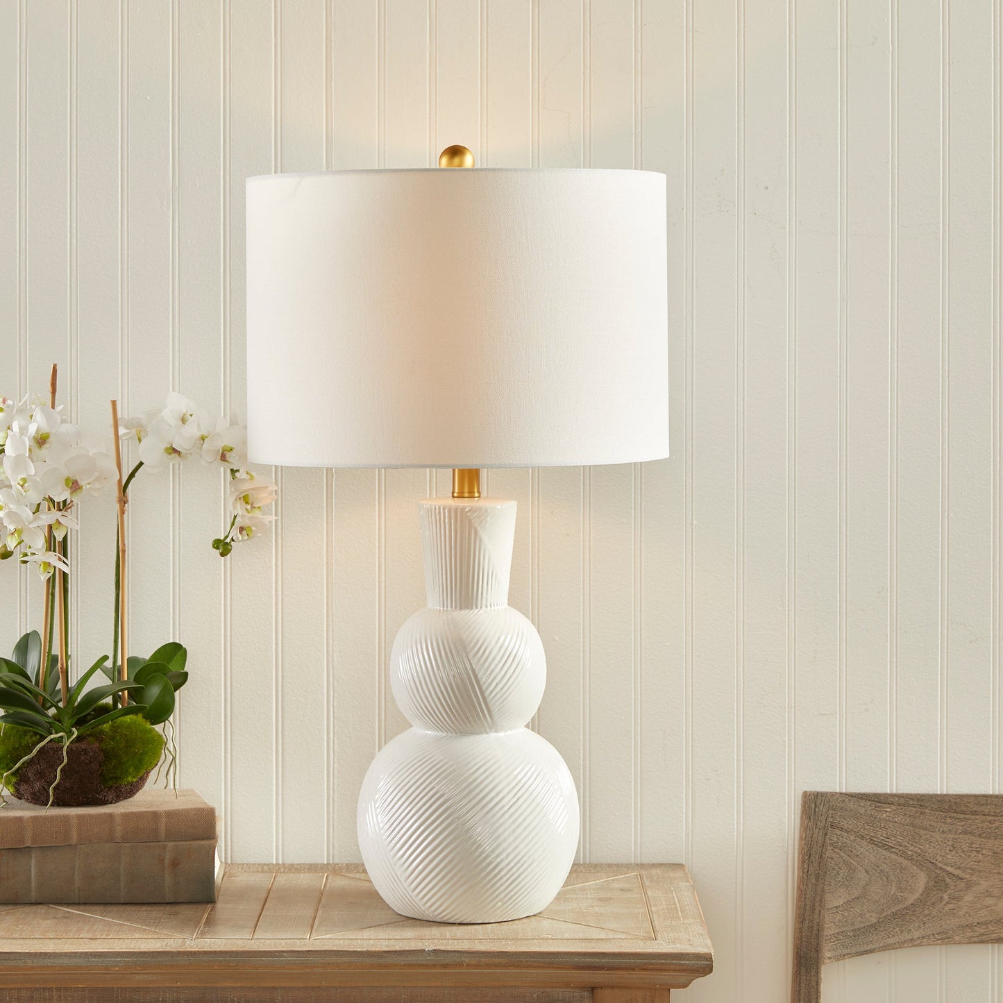 This traditional table lamp is crafted from clean white ceramic and features hand carved markings on the body, making it a perfect addition to any traditional setting. It is complemented by a white linen shade and a simple brass finial. Ideal for enhancing the ambiance of any living room or home decor.