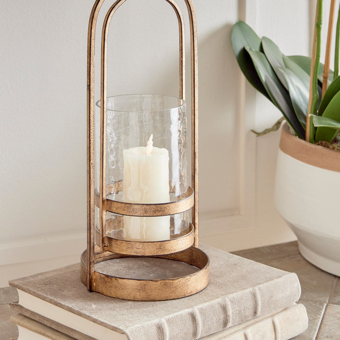 Crafted with precision from sturdy cast aluminum and adorned with exquisite hammered glass, the Amara Gold Hurricane Candle Holder Small showcases a perfect balance of lyrical form and substantial design. A decorative yet functional loop handle completes the look, making it a versatile addition to any transitional or modern setting.