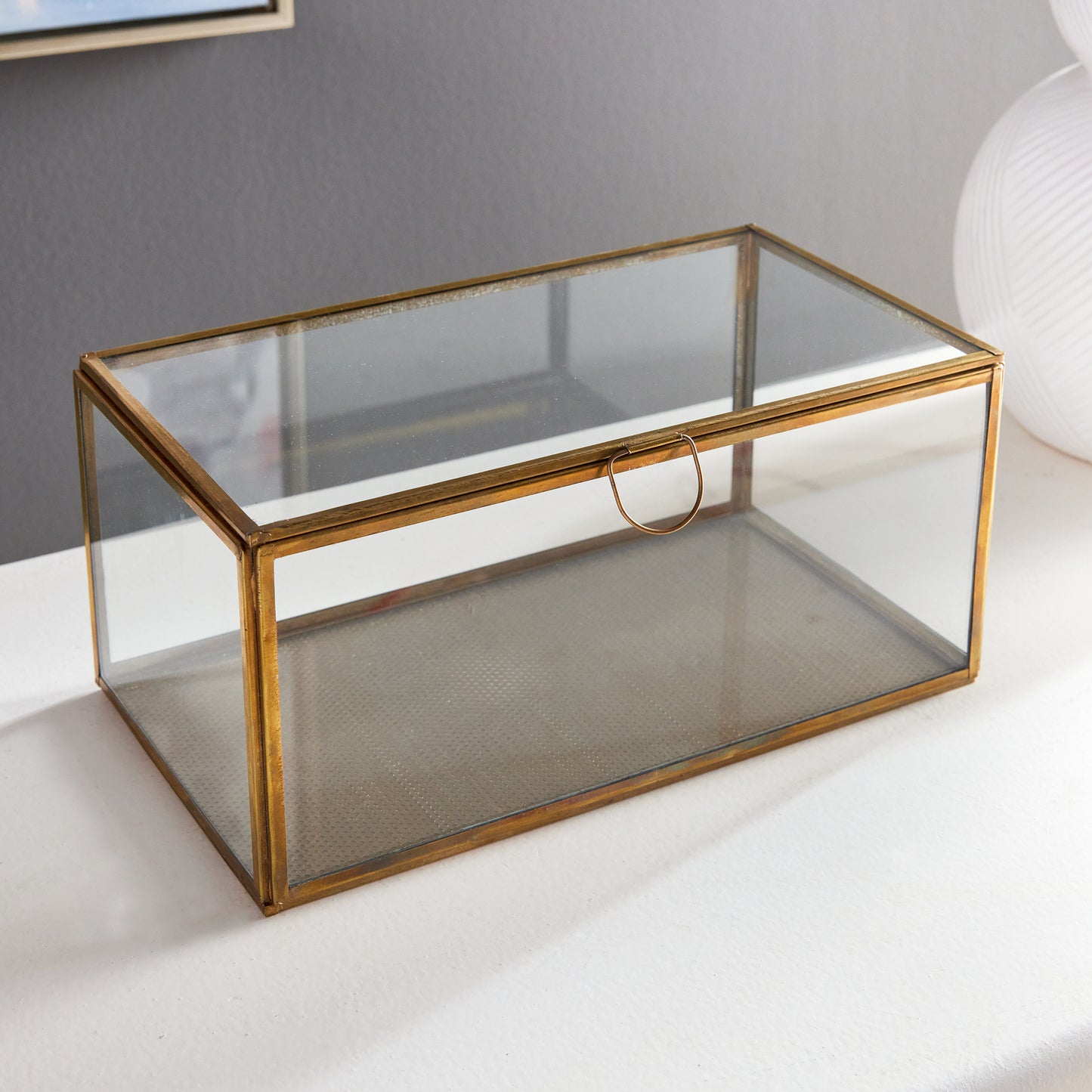 Expertly crafted with a combination of glass and brass, the Arwen Rectangular Antique Brass Display Box is designed to securely hold and protect your most cherished mementos. A sleek loop handle prevents fingerprints from marking the glass, keeping your keepsakes pristine.