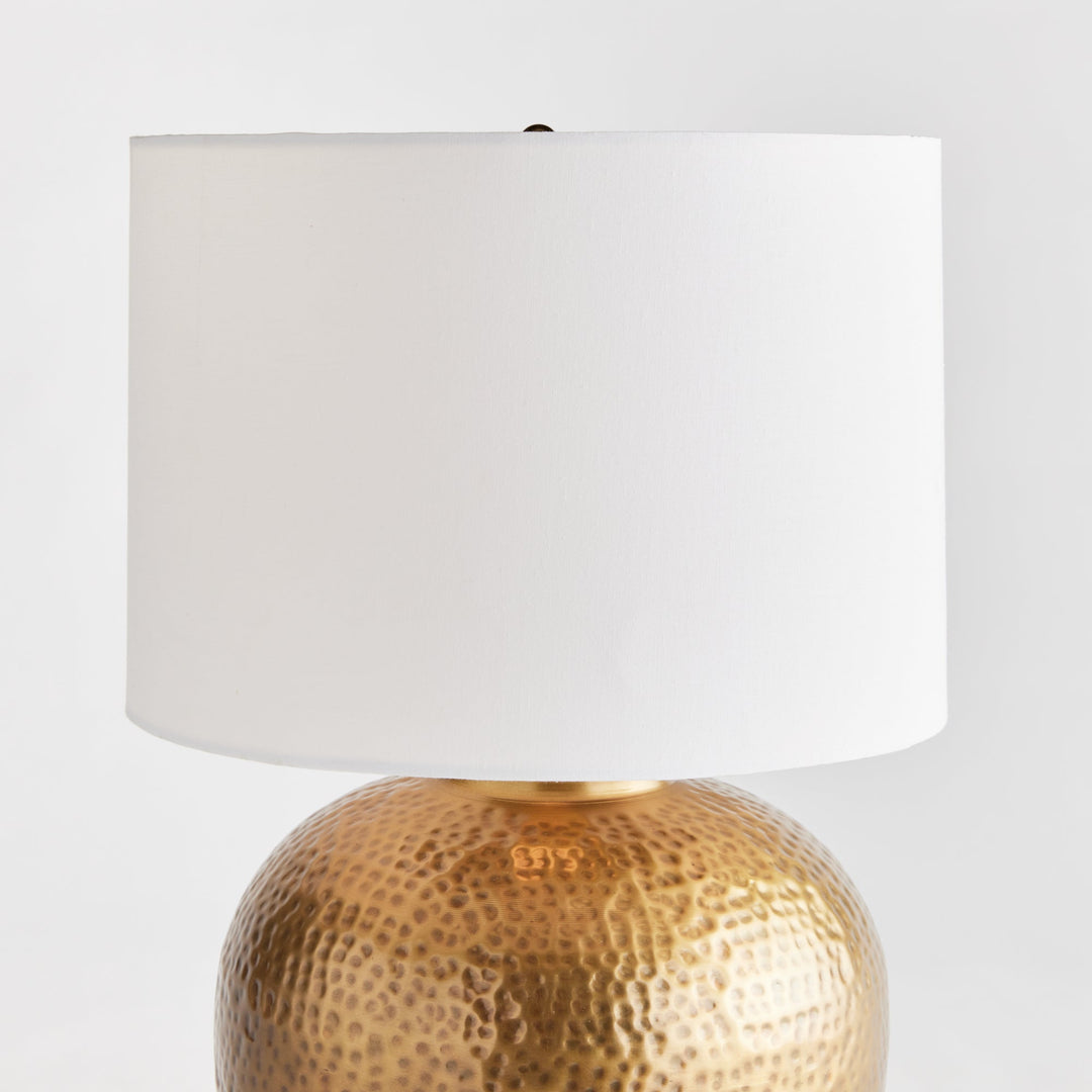 Enhance any room with the elegant style of Tara Gold &amp; White Table Lamp Small. The warm brass round base exudes a luxurious feel, while the crisp white linen drum shade offers a gentle radiance. Crafted from hammered iron and featuring unique handmade elements, this lamp is the perfect addition to any modern space.