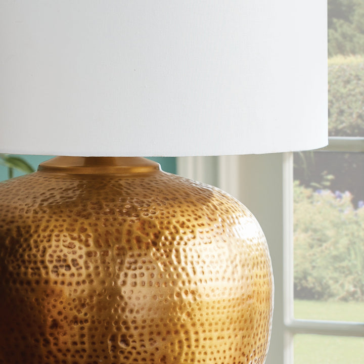 Crafted with a round base in warm brass and an impressive scale, the Tara Gold &amp; White Table Lamp serves as a handsome anchor for any room. Its clean white linen drum shade adds a soft touch and complements the bold design. Made from hammered iron, this one-of-a-kind lamp is sure to catch the eye and become a stunning focal point in your space.