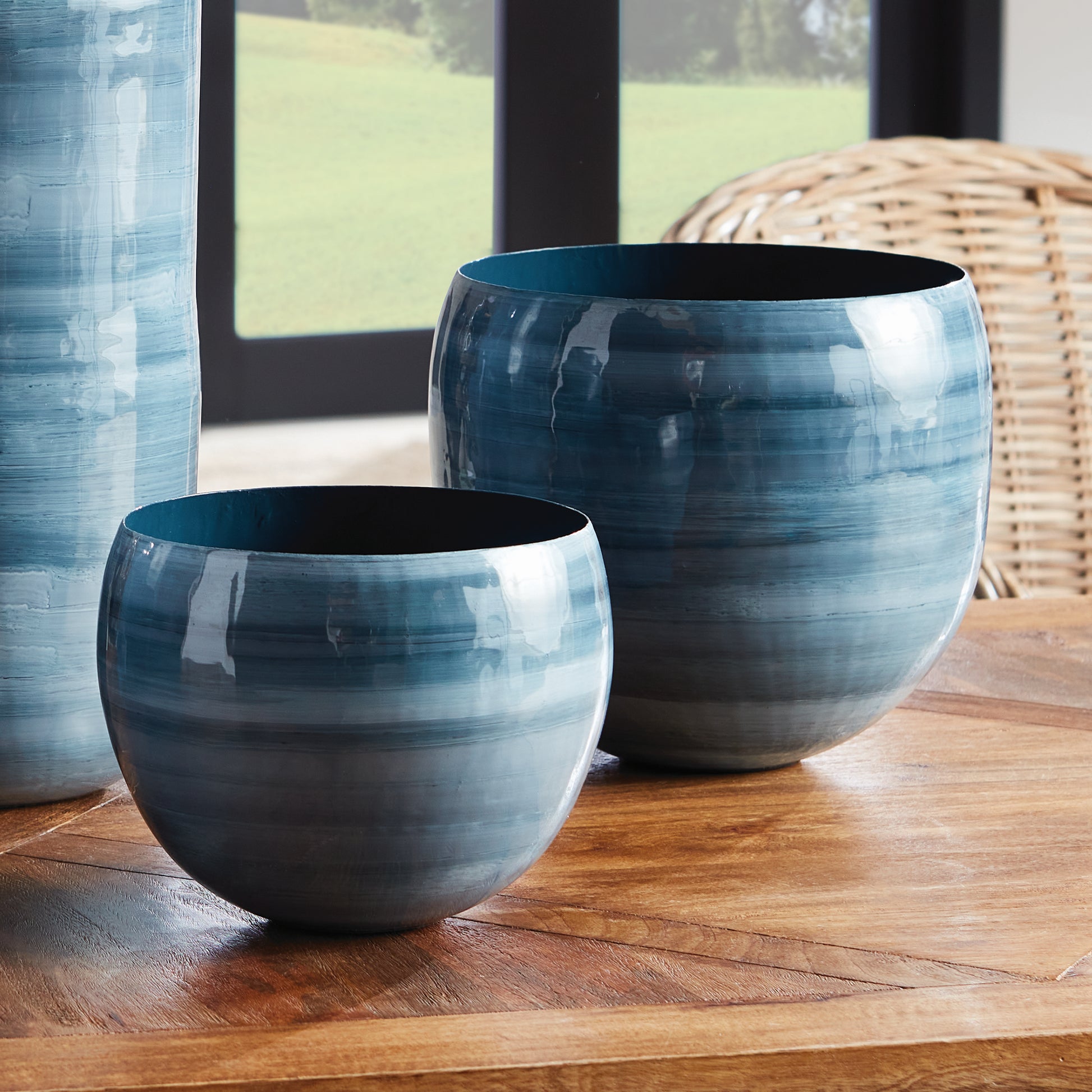 Crafted from lightweight enameled iron and meticulously hand-painted by experts, the Andrey Cache Planter Pots boast a refined, horizontal swirl design in a coastal-inspired blue gray palette. These cachepots serve as both functional pieces and unique works of art, adding a touch of modern sophistication to any space.