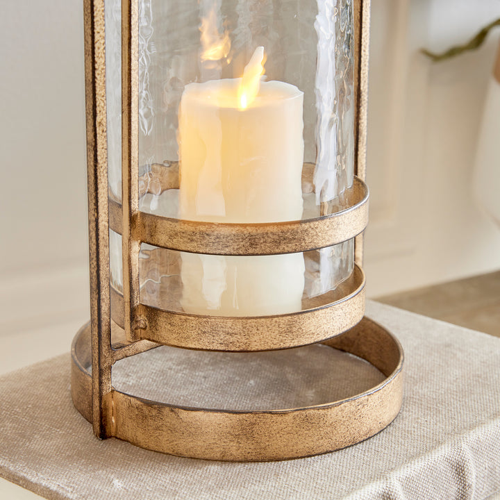 Crafted with precision from sturdy cast aluminum and adorned with exquisite hammered glass, the Amara Gold Hurricane Candle Holder Small showcases a perfect balance of lyrical form and substantial design. A decorative yet functional loop handle completes the look, making it a versatile addition to any transitional or modern setting.