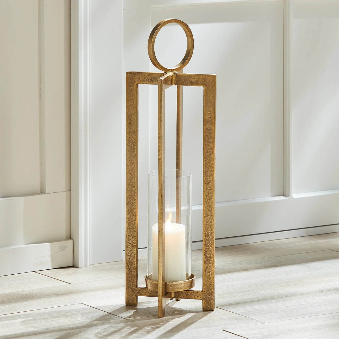 Elevate your space with the Asola Gold Hurricane Lantern Small. Exuding modern sophistication, this lantern boasts a striking form and impressive scale. With a footed detail that showcases your candle in style and a large round handle for effortless transportation, it's the perfect addition to mantles, front entries, and any other area in need of a touch of elegance.