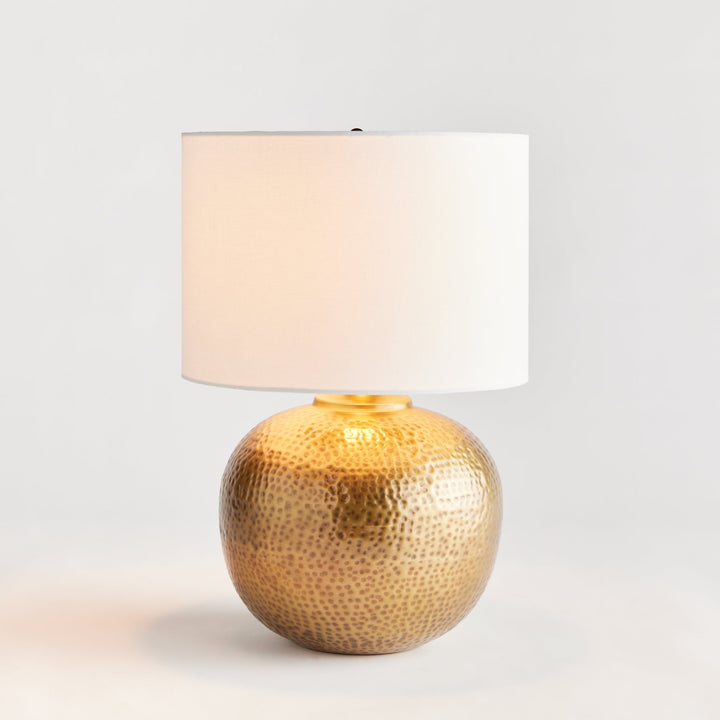 Enhance any room with the elegant style of Tara Gold &amp; White Table Lamp Small. The warm brass round base exudes a luxurious feel, while the crisp white linen drum shade offers a gentle radiance. Crafted from hammered iron and featuring unique handmade elements, this lamp is the perfect addition to any modern space.