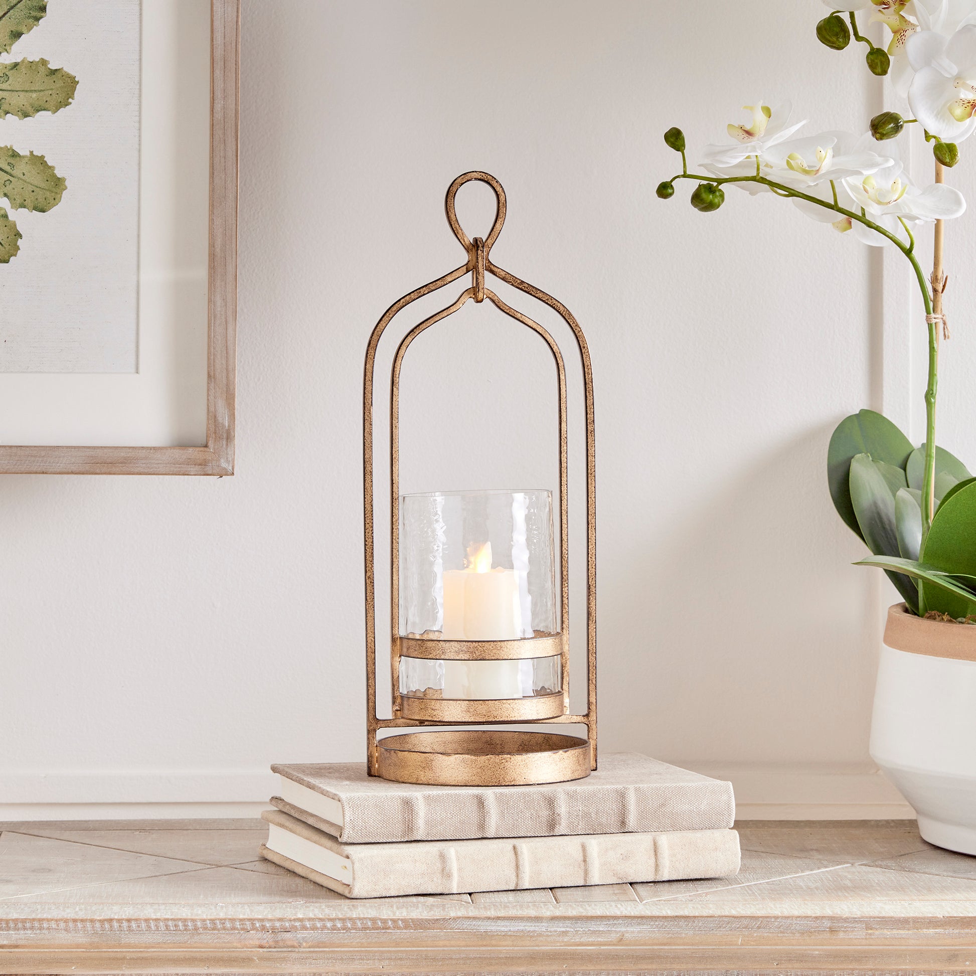 Crafted with precision from sturdy cast aluminum and adorned with exquisite hammered glass, the Amara Gold Hurricane Candle Holder Small showcases a perfect balance of lyrical form and substantial design. A decorative yet functional loop handle completes the look, making it a versatile addition to any transitional or modern setting.