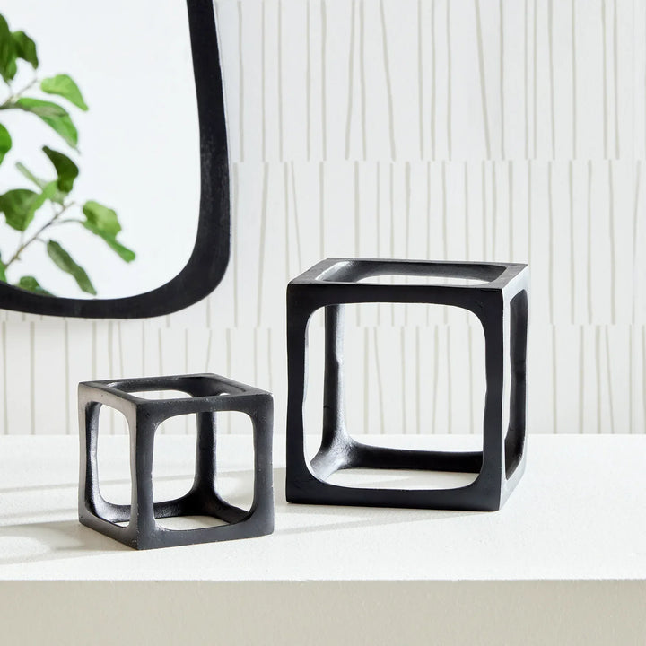 Two black cube-shaped decorative sculptures on a light surface with a plant in the background.