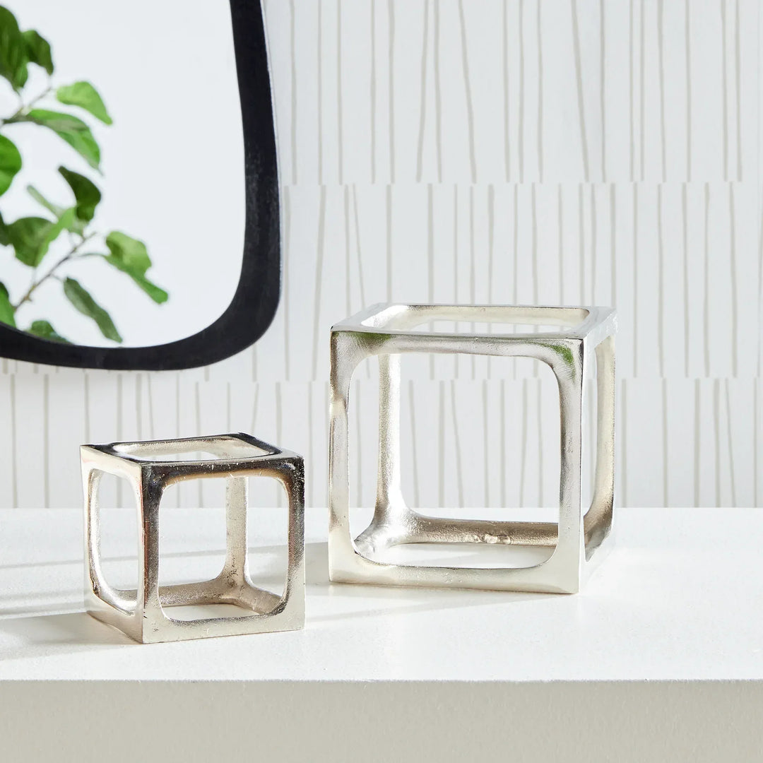 Two metallic square sculptures on a white surface with a blurred plant in the background