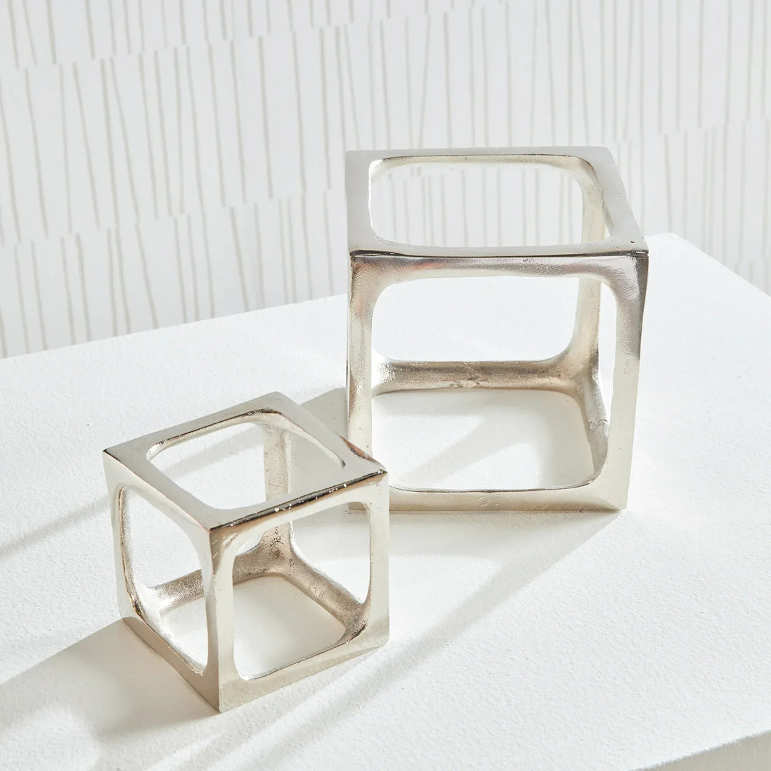 Two metallic cube-shaped sculptures on a white surface with a light gray textured background