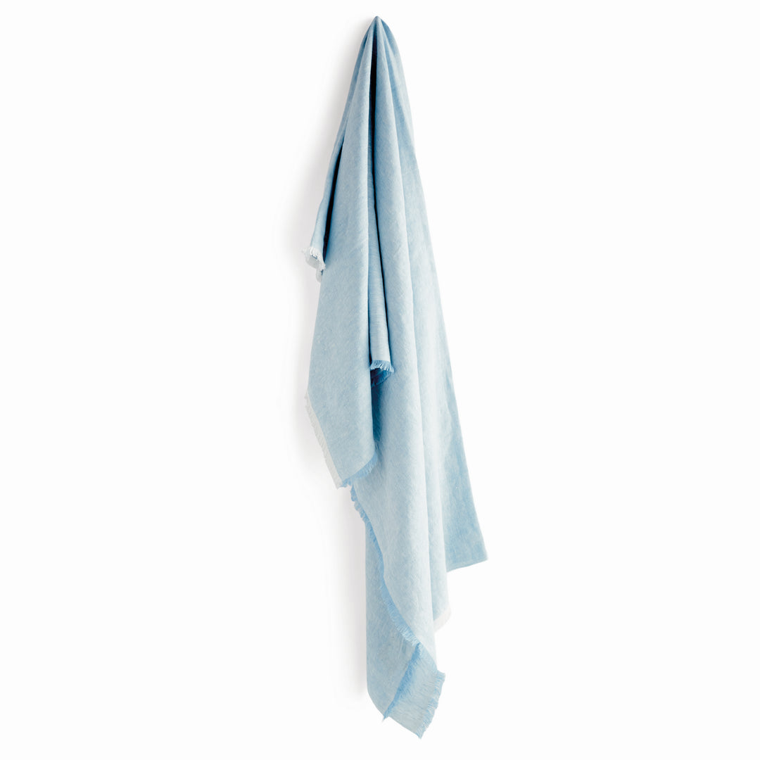 Light blue European linen throw blanket hanging elegantly against a white wall, showcasing its drape and soft texture.