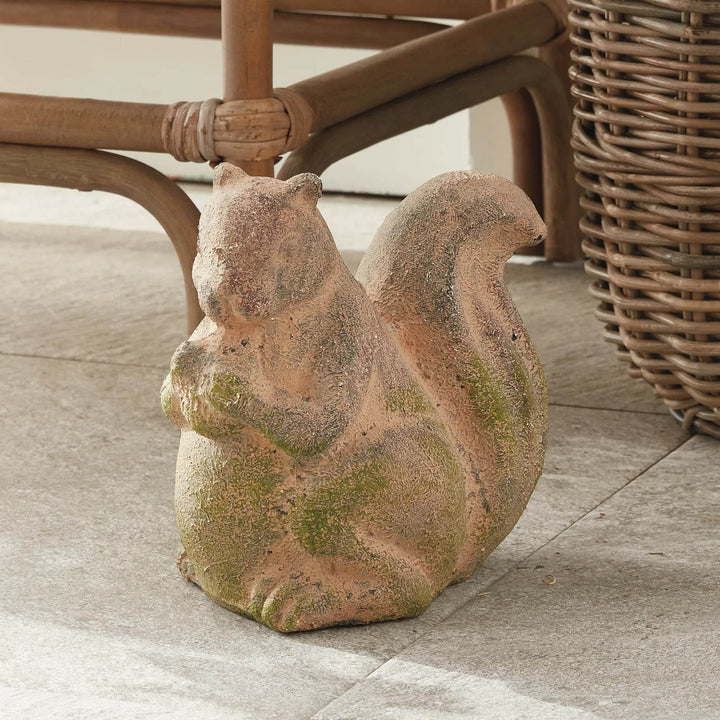 Weathered terracotta squirrel sculpture with mossy finish, placed outdoors next to wicker furniture and natural stone flooring.

