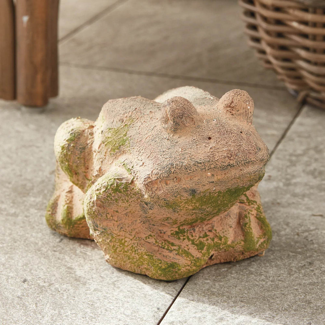 Moss terracotta frog sculpture with weathered finish, styled outdoors on stone patio next to wicker and wood furniture.

