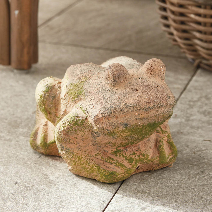 Moss terracotta frog sculpture with weathered finish, styled outdoors on stone patio next to wicker and wood furniture.

