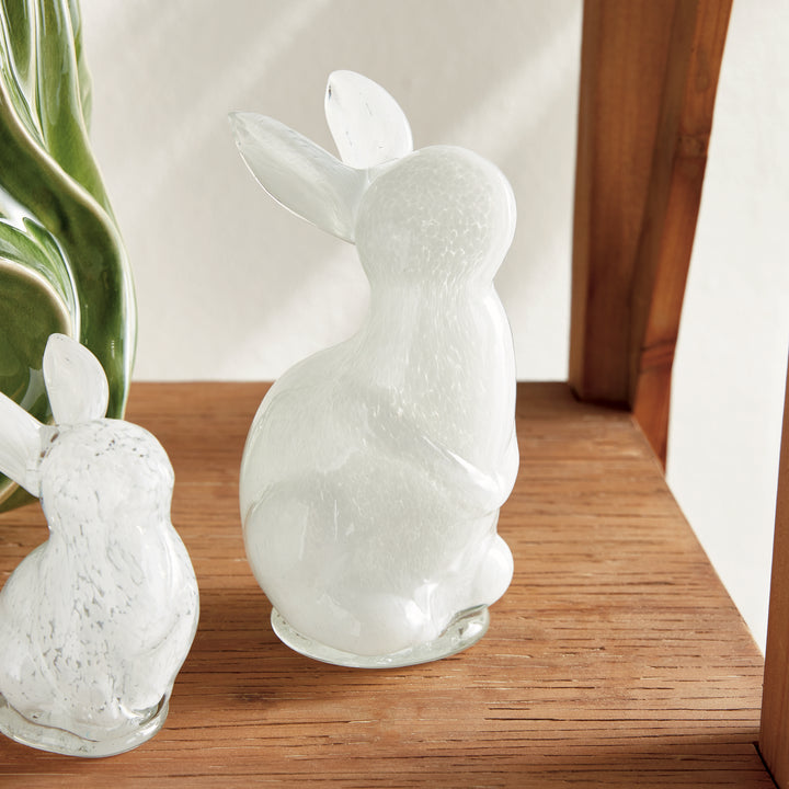 Two hand blown glass rabbits on a wooden surface with a plant in the background.