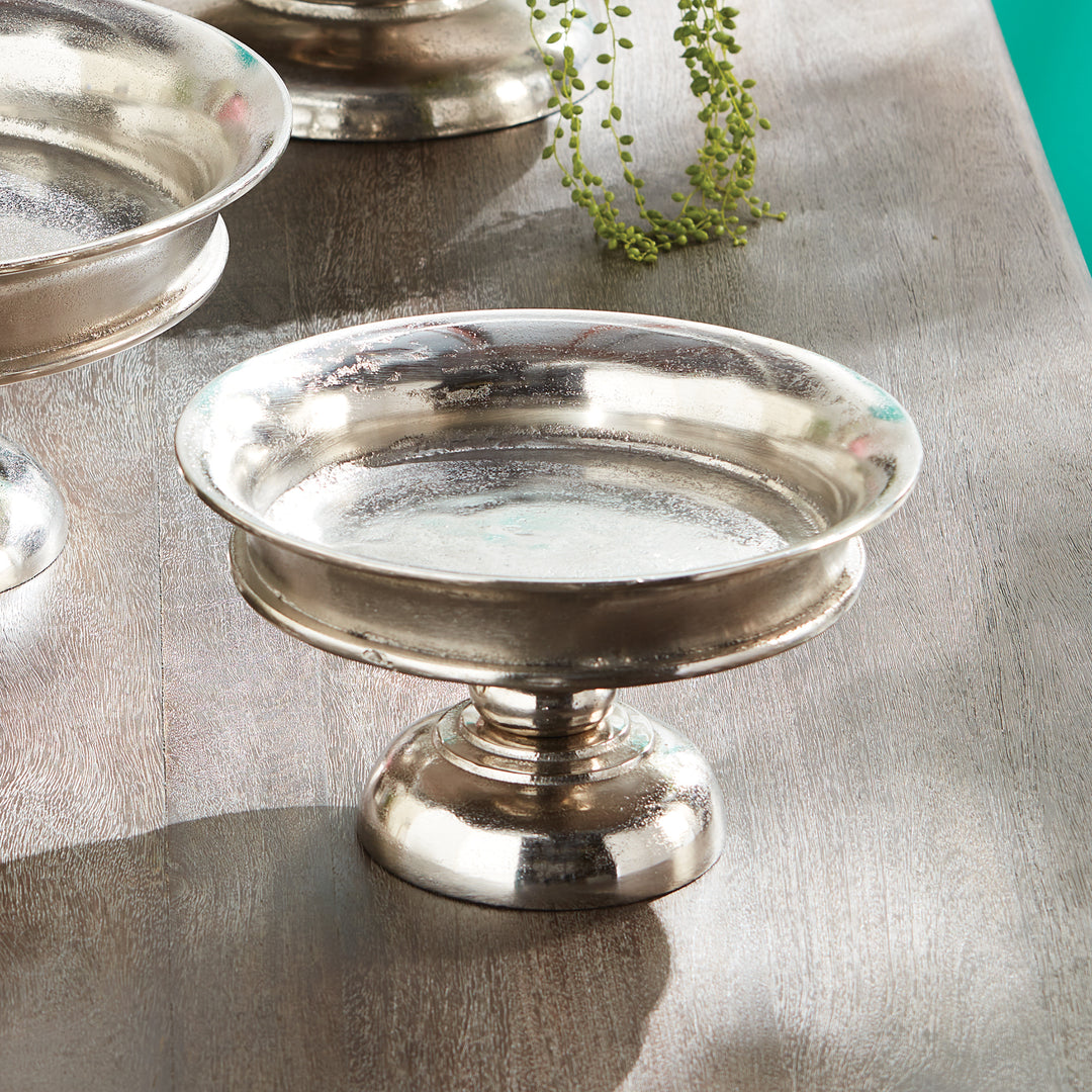 Silver pedestal dish on a reflective surface with a plant in the background