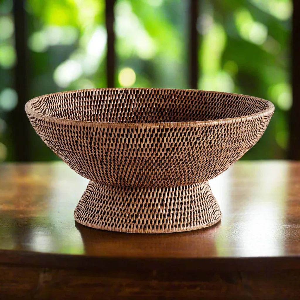 Warm brown woven rattan pedestal offering bowl on a polished wooden table with a softly blurred green backdrop, Transitional