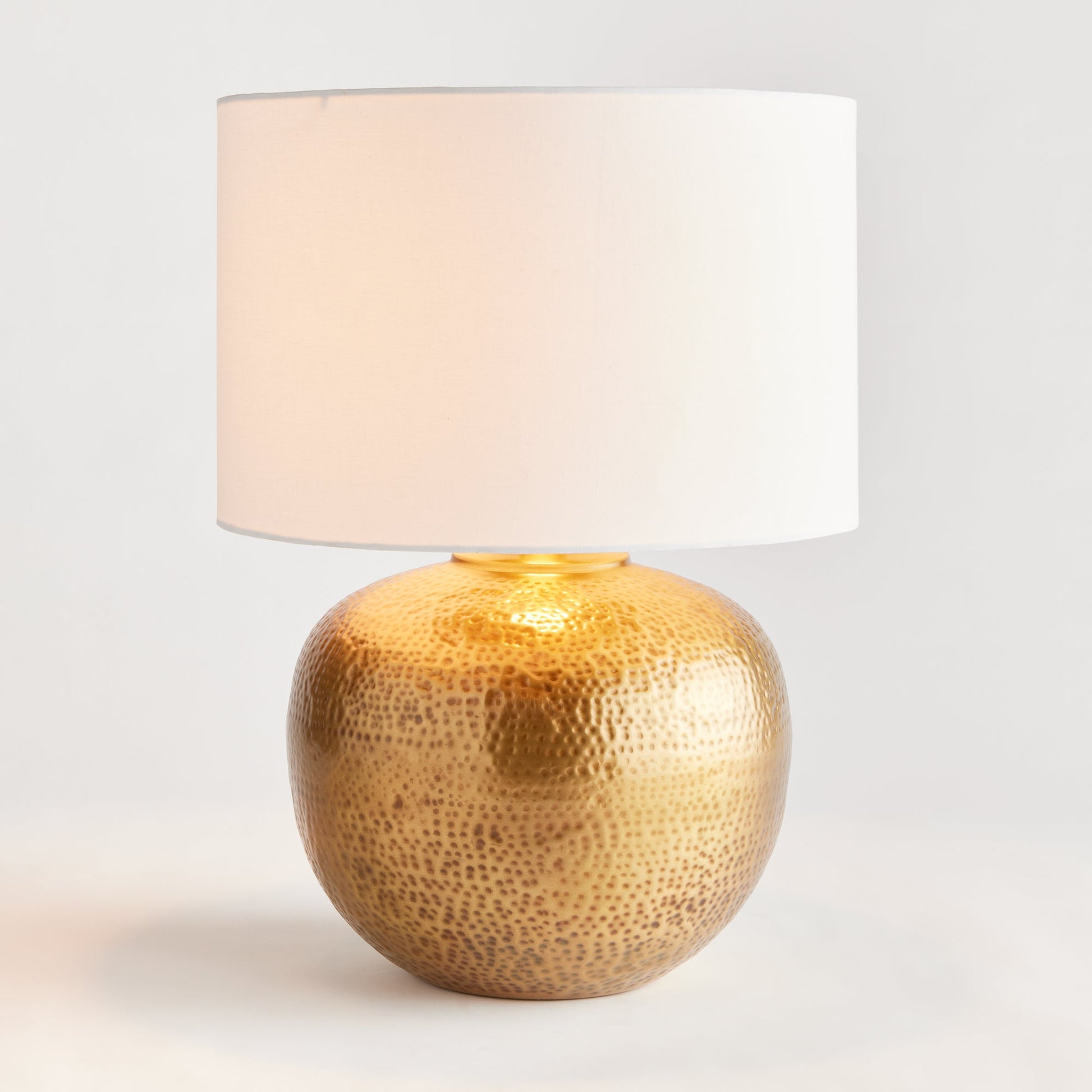 Crafted with a round base in warm brass and an impressive scale, the Tara Gold &amp; White Table Lamp serves as a handsome anchor for any room. Its clean white linen drum shade adds a soft touch and complements the bold design. Made from hammered iron, this one-of-a-kind lamp is sure to catch the eye and become a stunning focal point in your space.