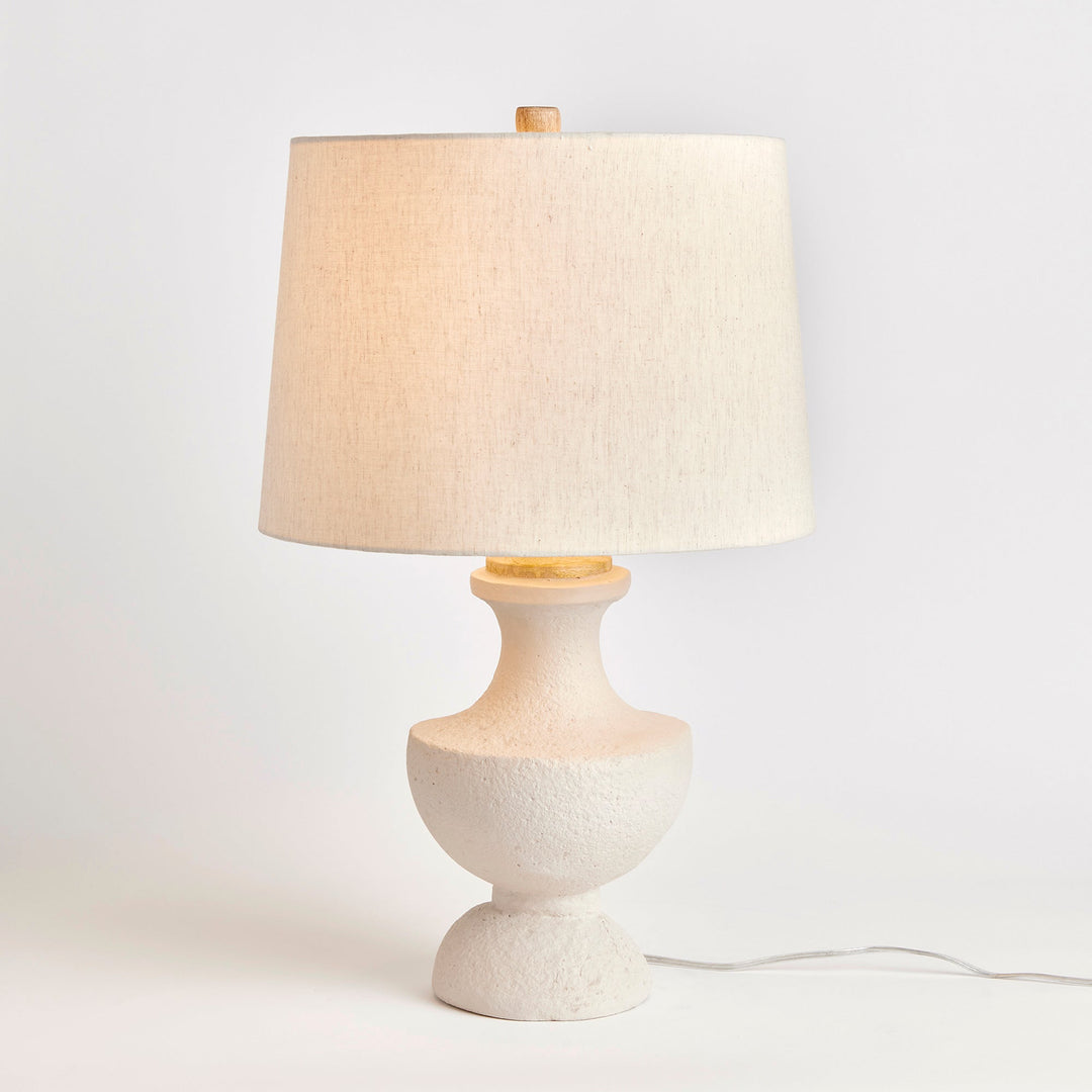 Constructed with a combination of Eco-mix and composite materials, this lamp base boasts durability with its hardened agents. Its warm white finish showcases the material's composition through a textured and pitted surface. Featuring a linen tapered shade and classic silhouette, this diminutive lamp is ideal for a bedside table or desk in need of illumination.