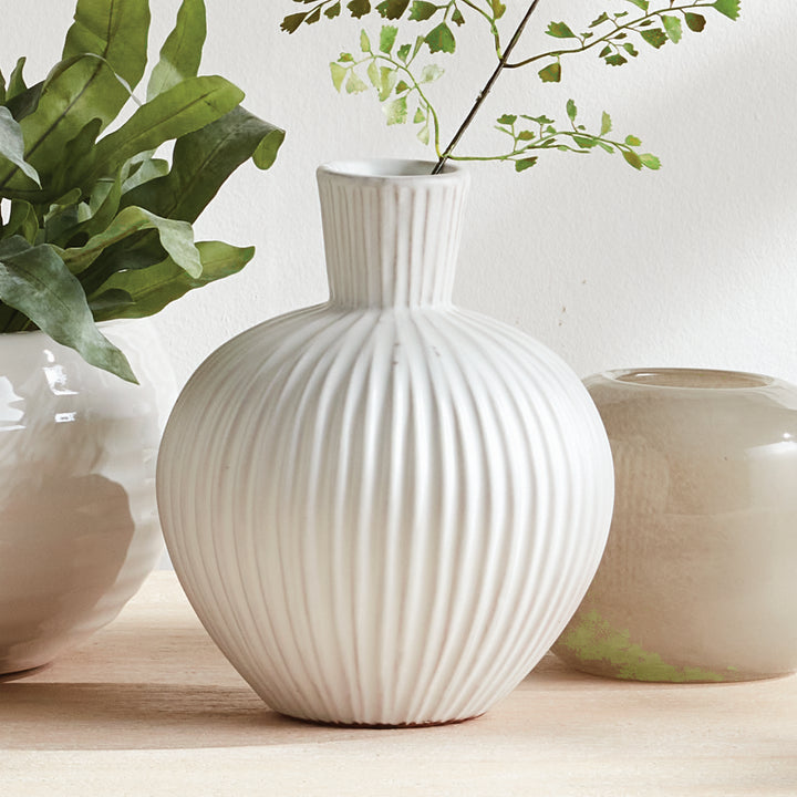 White ribbed terracotta bottle vase with green stems beside other pottery, showing its round body and tall neck. Transitional