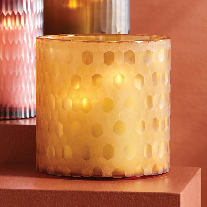Amber glass hurricane styled on shelf with glowing candlelight and etched texture, shown with coordinating modern glass vessels – Modern Glam.

