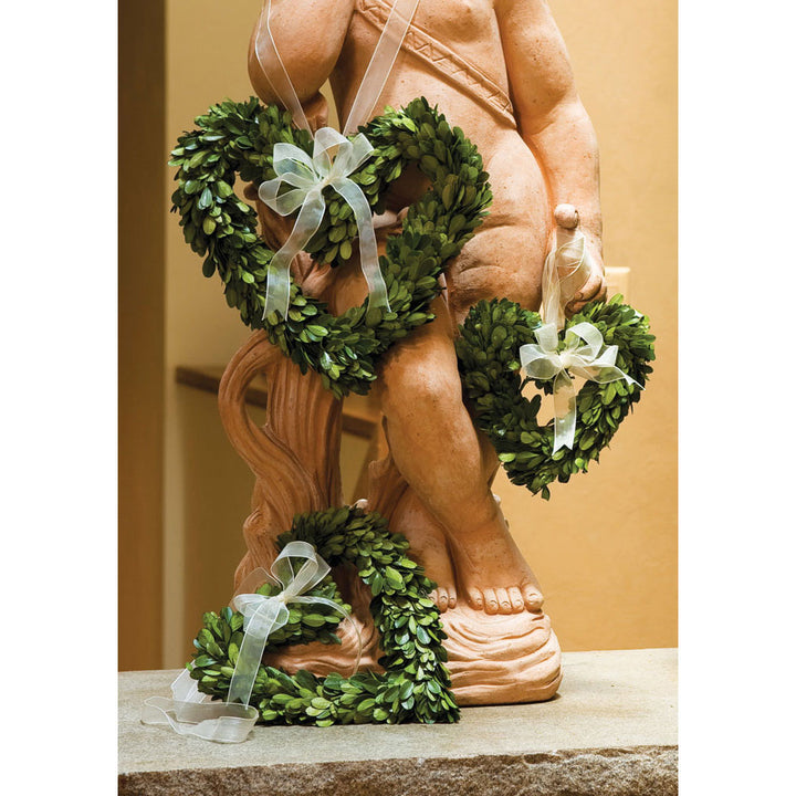 Amity preserved boxwood heart wreaths with pale ribbons arranged around a carved figure in a warm interior. Transitional