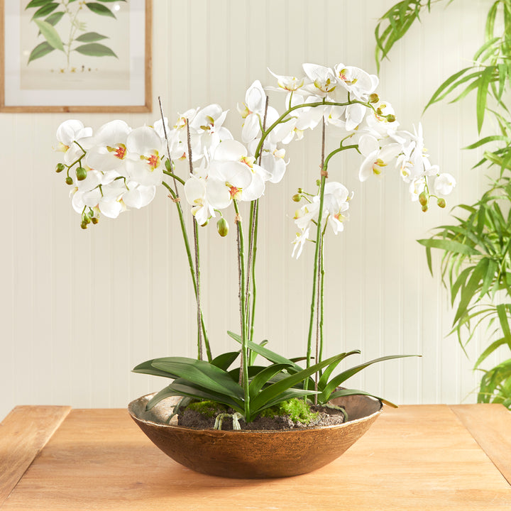 Antique brass decorative bowl styled with tall white orchids on a wooden dining table in a bright interior, Modern Organic