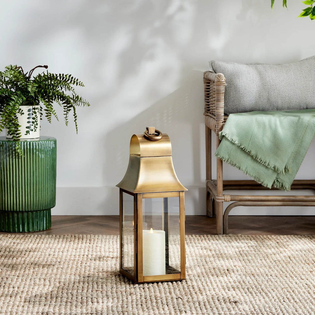 Tall brass lantern styled beside woven bench and green ceramic planter, shown with LED candle for indoor décor – Transitional Home Accent