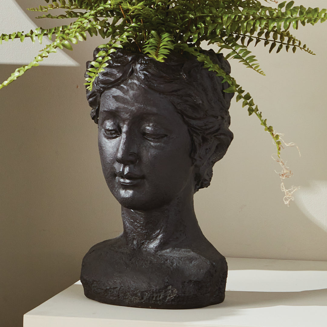 Antique black female bust planter styled with fern foliage above its sculpted hair, standing on a pale surface. Transitional