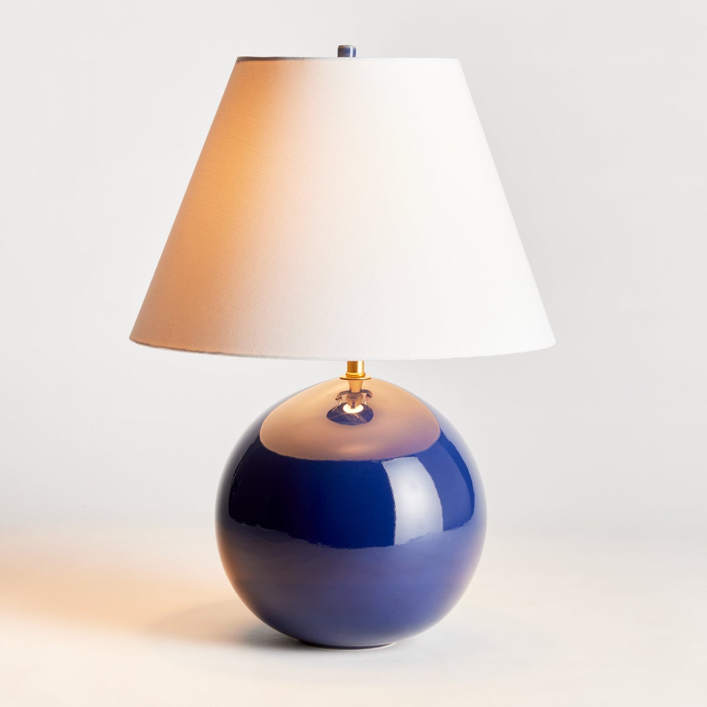 With a perfectly rounded ceramic body and a deep blue high gloss glaze, this table lamp adds a pop of color to any setting. Topped off with a bright white tapered shade, it makes a lovely accent for bedside table, side table or desk. Expertly crafted with a rounded ceramic body and a stunning high gloss blue glaze, this lamp is sure to elevate any space. Paired with a sleek and bright white tapered shade, it's the perfect addition to your bedside table, side table, or desk.