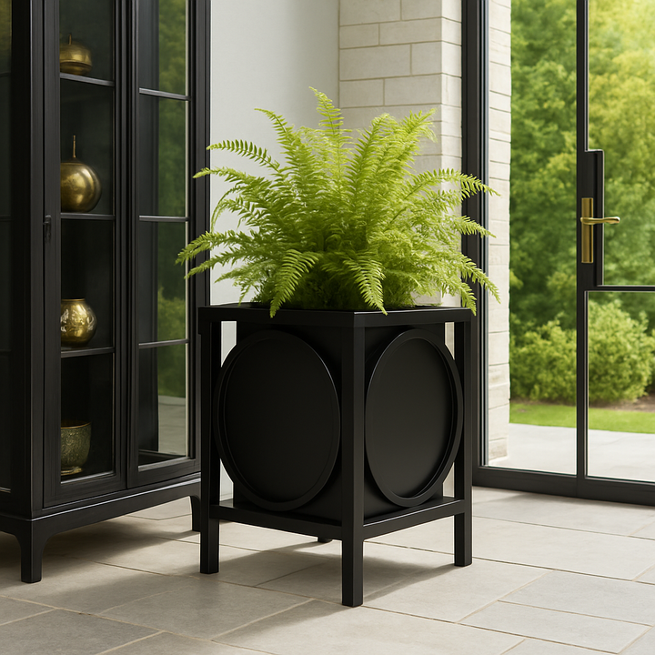 Matte black iron planter with circular detail, styled beside a glass cabinet on a tiled patio with heritage accents and lush garden backdrop.