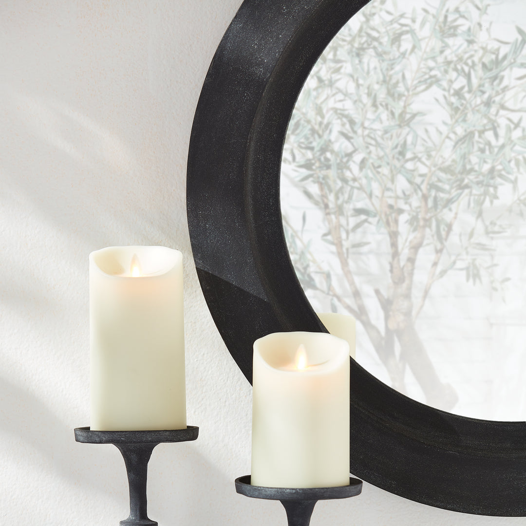Close-up of black frame round wall mirror showing soft candle glow and reflected olive tree in clean modern interior.

