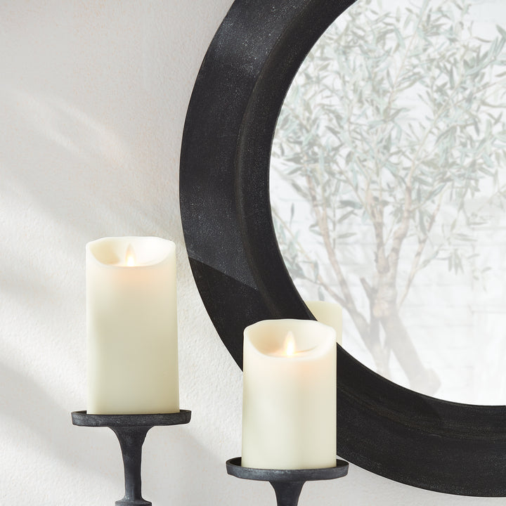 Close-up of black frame round wall mirror showing soft candle glow and reflected olive tree in clean modern interior.

