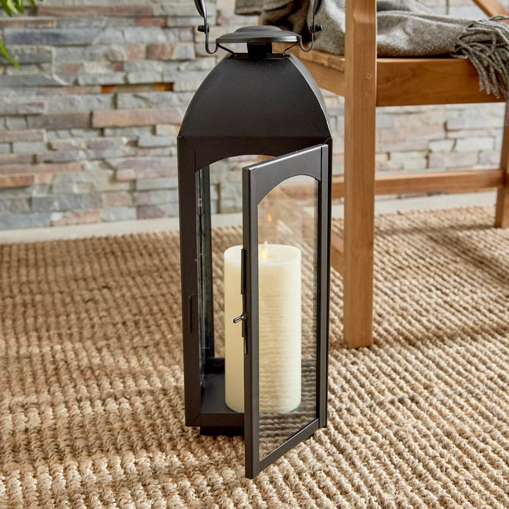 Black outdoor lantern with lit pillar candle on woven rug; glass panes and arched door give evening ambiance – Transitional Home Decor