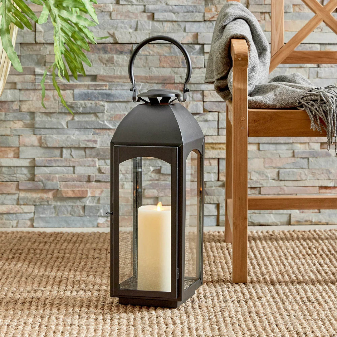 Large black outdoor lantern with pillar candle styled by chair and stone wall, woven rug; glass panes visible – Transitional Styling.