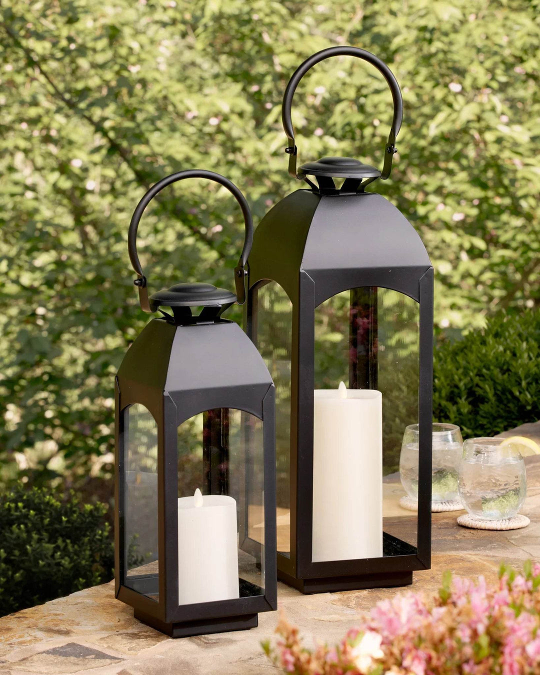 Pair of black outdoor lanterns glowing in garden at dusk; glass panes, iron frames and ring handles by plants – Transitional Yard Decor