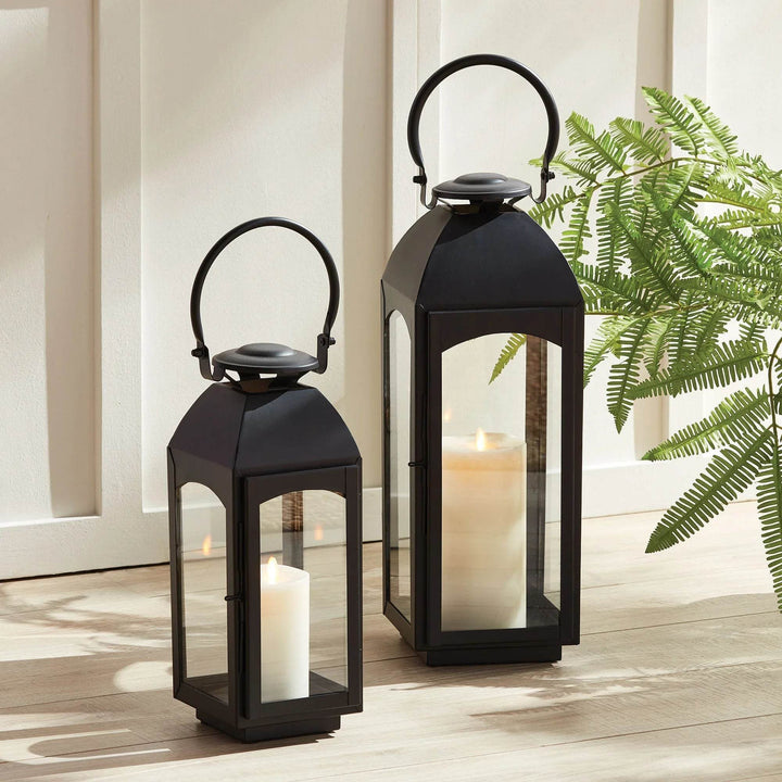 Two black outdoor lanterns, small and large, styled by a sunlit wall; glass panes and pillar candles add glow – Transitional Outdoor Lanterns.