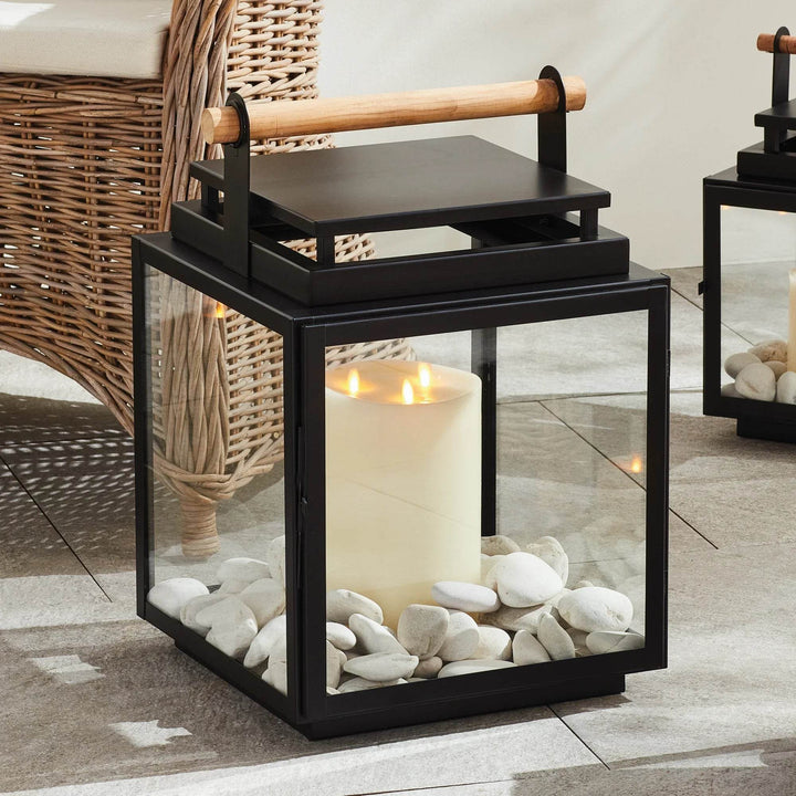 Large black outdoor lantern with teak handle shown outdoors on patio, styled with pillar candle and white stones – Modern Farmhouse.