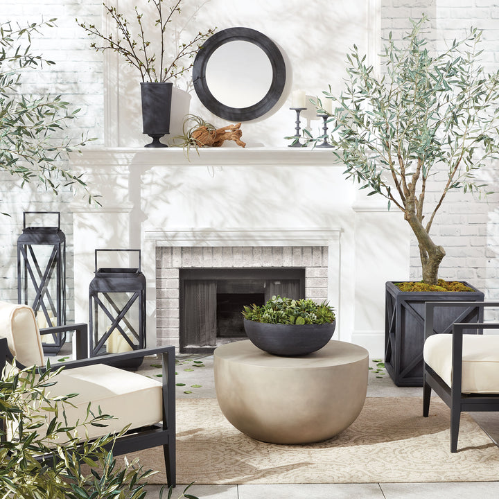 Black round wall mirror styled above white fireplace with olive trees, lanterns, and outdoor seating in a modern garden setting.
