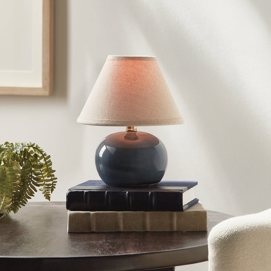 Blue ceramic mini table lamp lit on stacked books beside a fern, with a glowing cream linen shade - Coastal Transitional