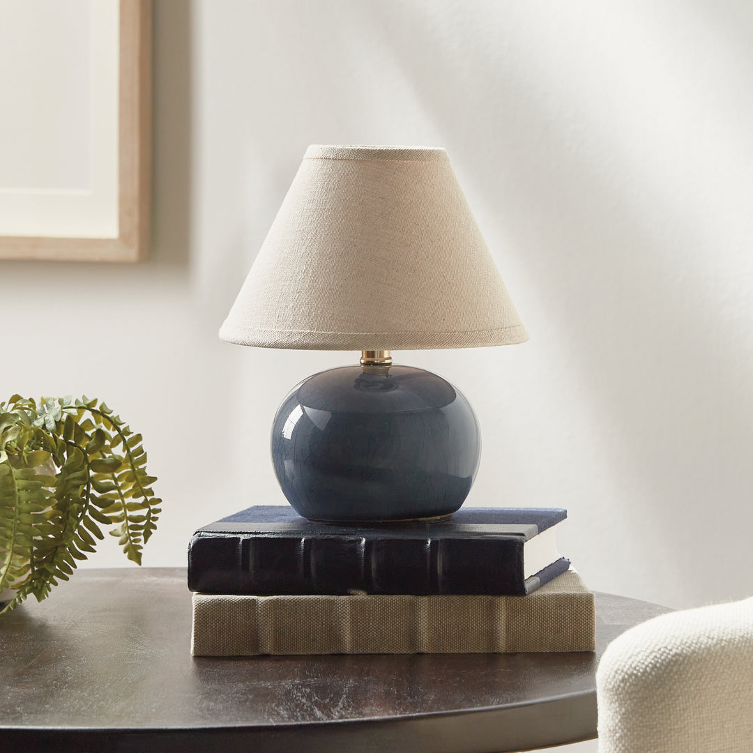 Blue ceramic mini table lamp on stacked books beside a fern, with a rounded base and linen lampshade - Coastal Transitional
