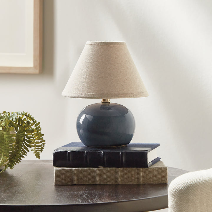 Blue ceramic mini table lamp on stacked books beside a fern, with a rounded base and linen lampshade - Coastal Transitional