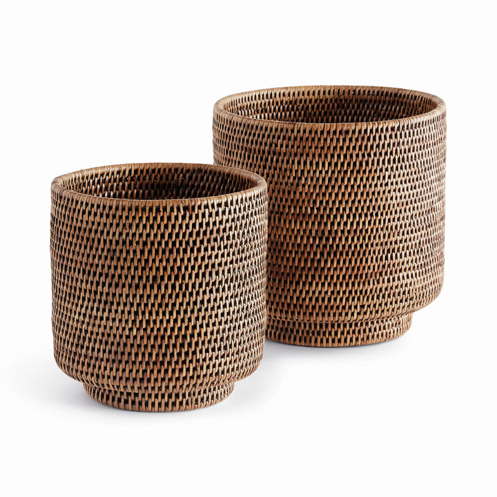 Brown rattan cachepots in two sizes with cylindrical woven sides and small round feet, shown on white - Coastal Transitional